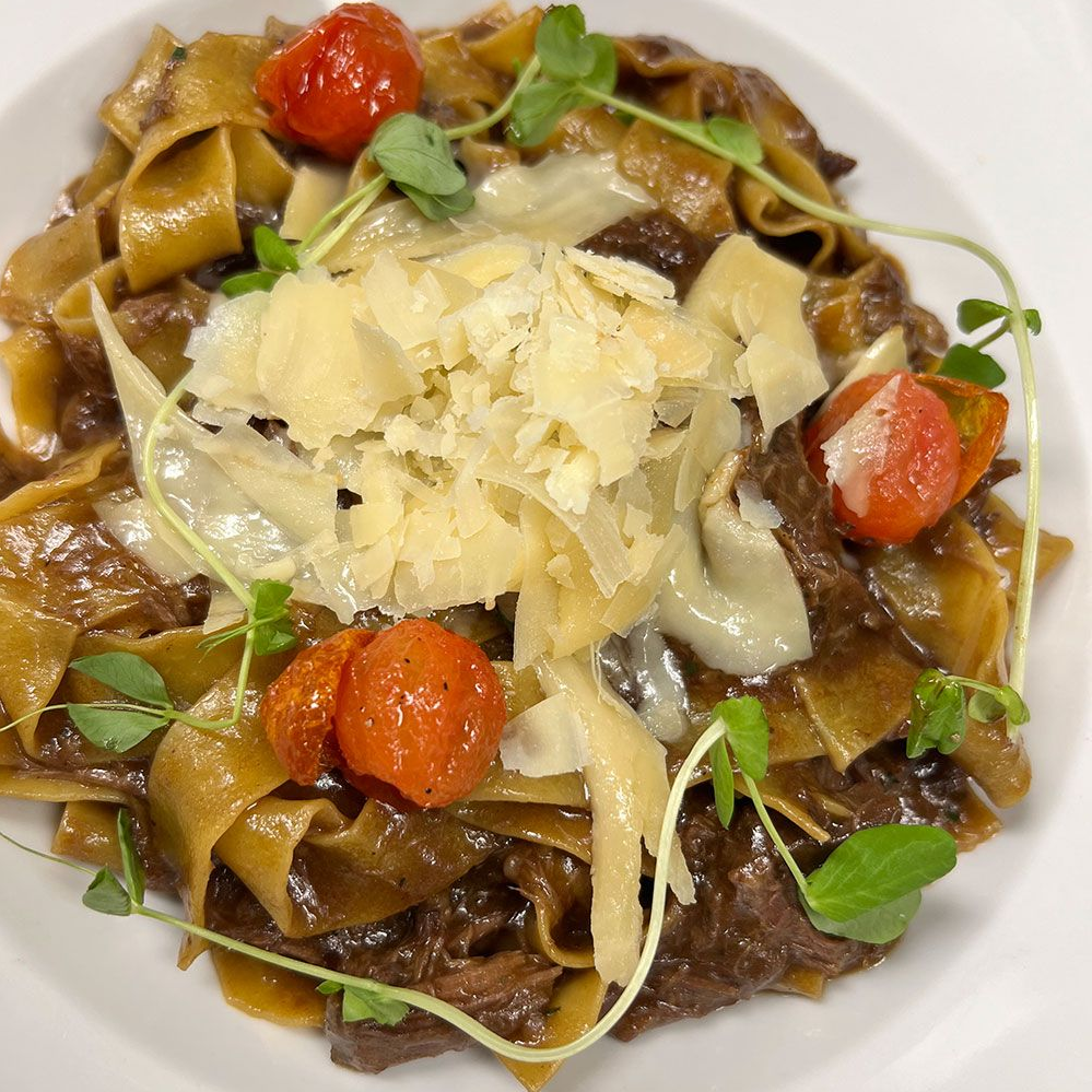 A serving of pappardelle pasta with beef ragu, topped with shaved parmesan, cherry tomatoes, and microgreens.