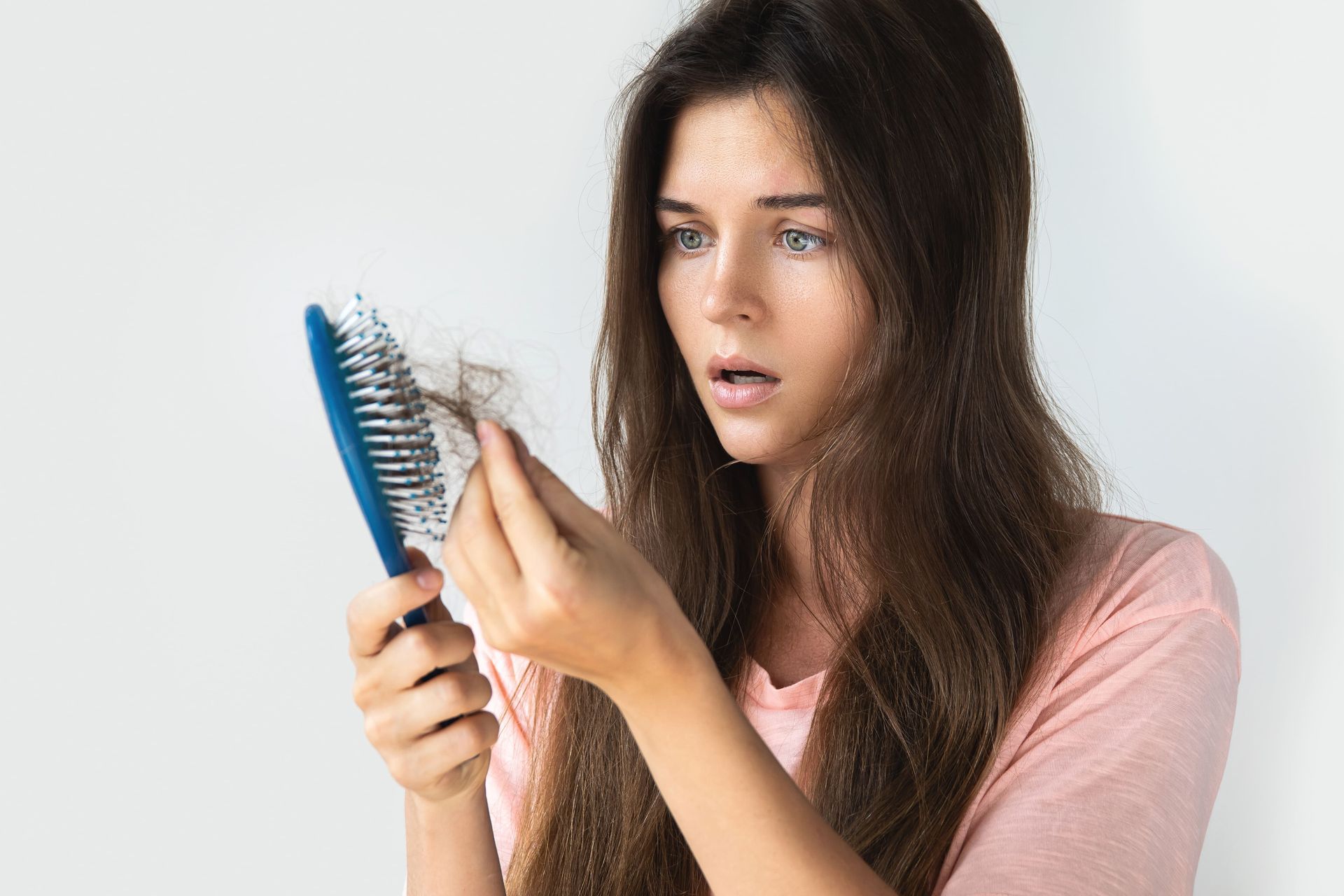 Mulher segurando uma escova de cabelo e examinando os fios emaranhados, com expressão de surpresa contra um fundo branco.
