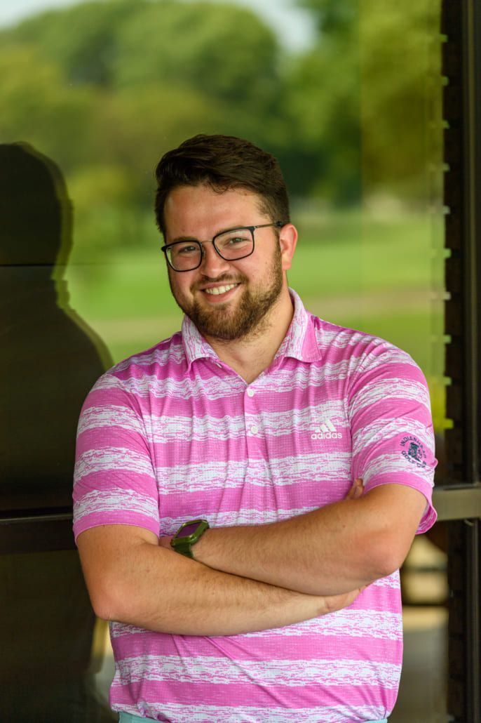 A man wearing glasses and a pink striped shirt is standing with his arms crossed.