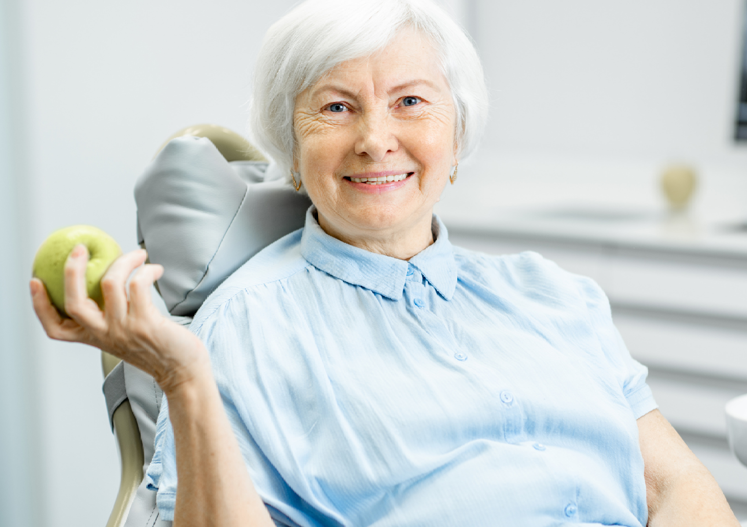 An elderly woman is sitting in a dental chair holding an apple.
