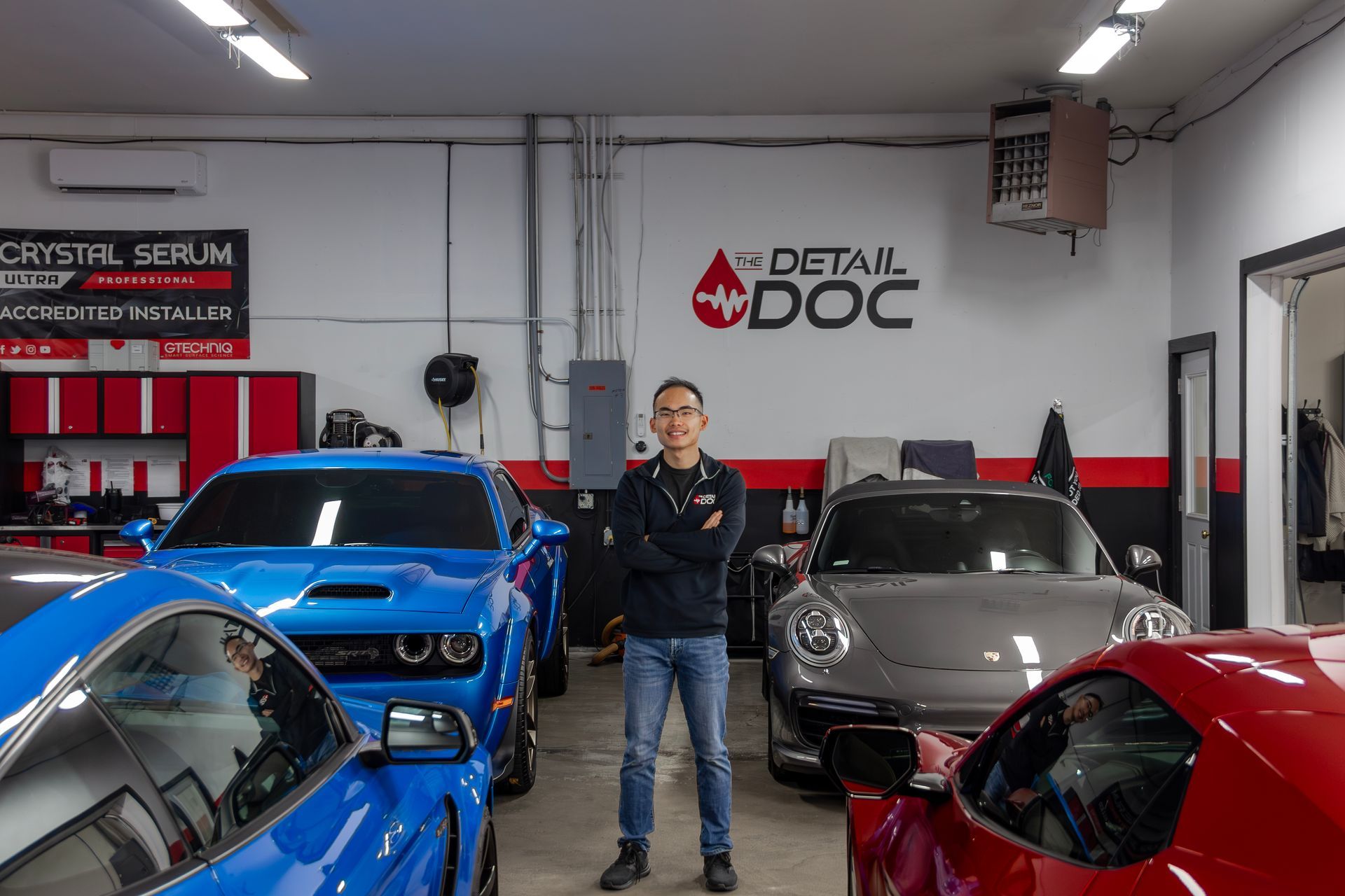 Man in auto detailing shop, arms crossed, stands between four cars: blue Mustang, blue Dodge, silver Porsche, and red Corvette.