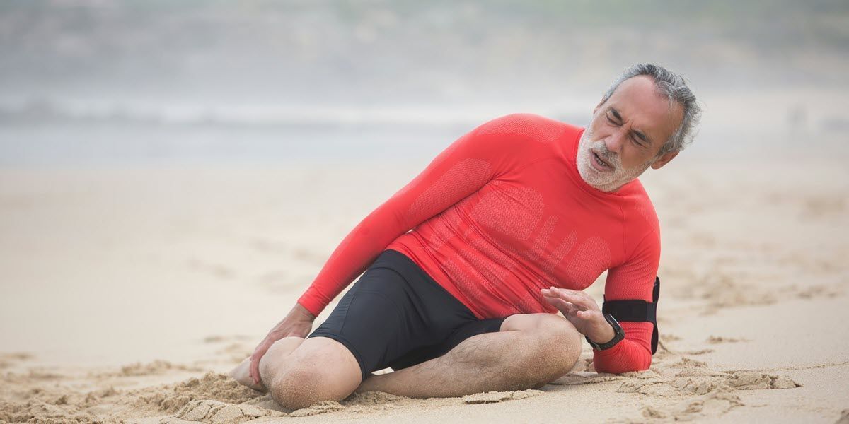 Man in a red swim shirt lying on the beach, leaning on one arm and holding his ankle.
