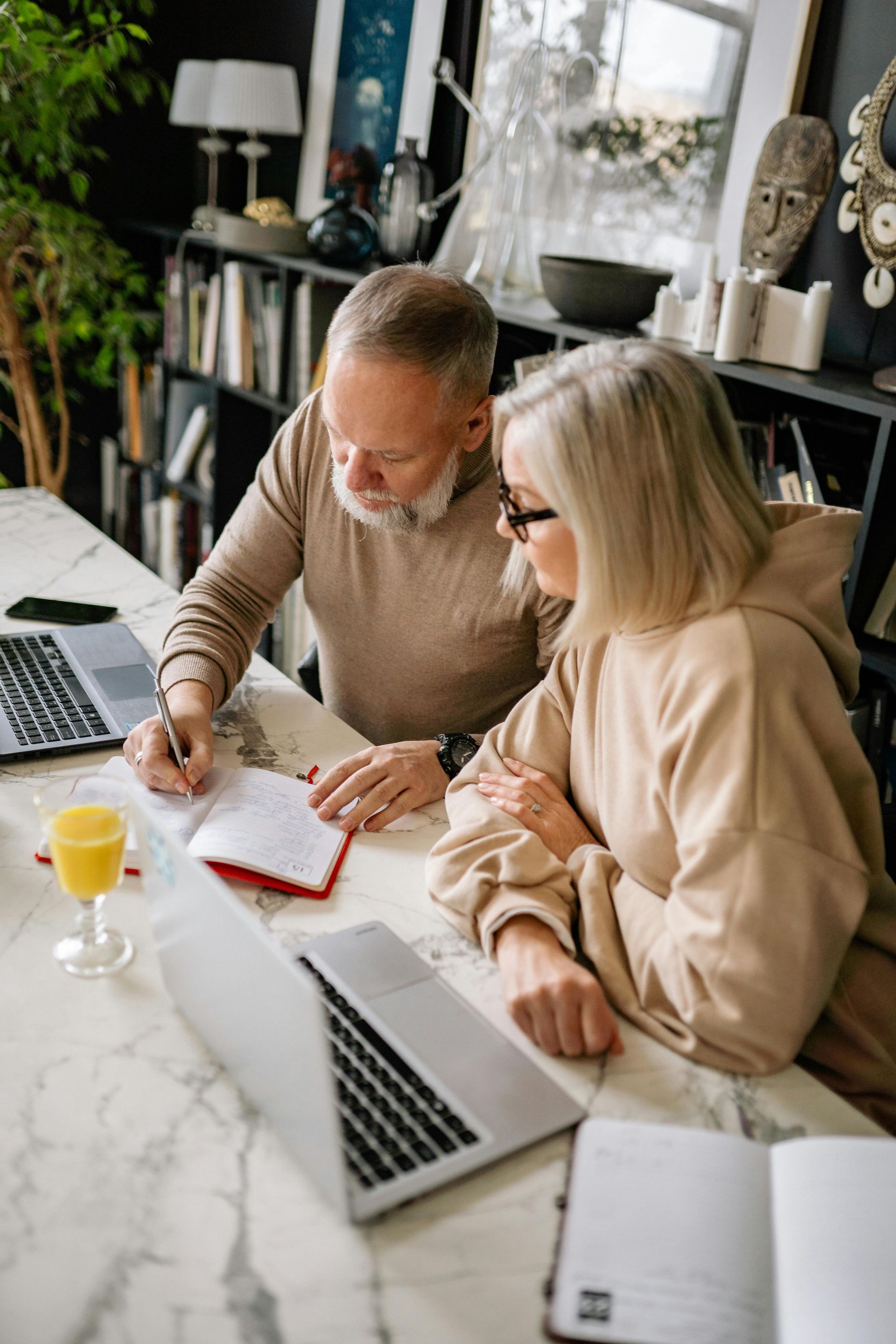 A man and a woman are sitting at a table with a laptop writing notes in a notebook.