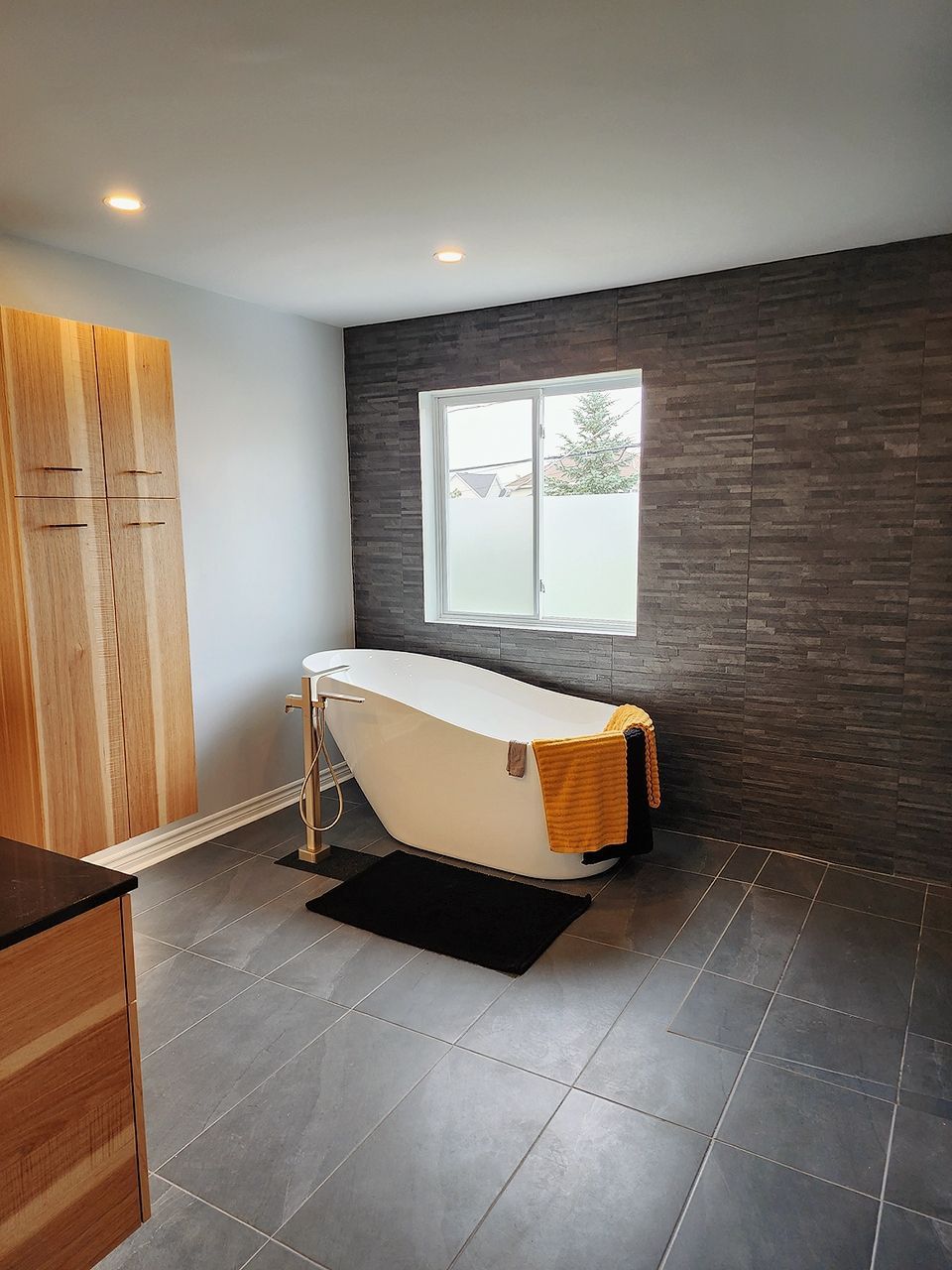 Salle de bain moderne avec baignoire autoportante, mur d'accent en carrelage foncé, armoires en bois et sol en carrelage.
