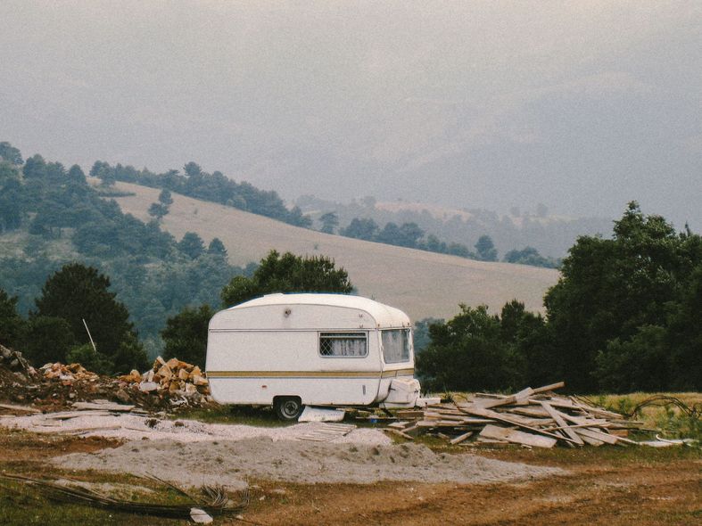 White camper trailer on a gravel lot beside stacked wood, with wooded hills in a hazy background.