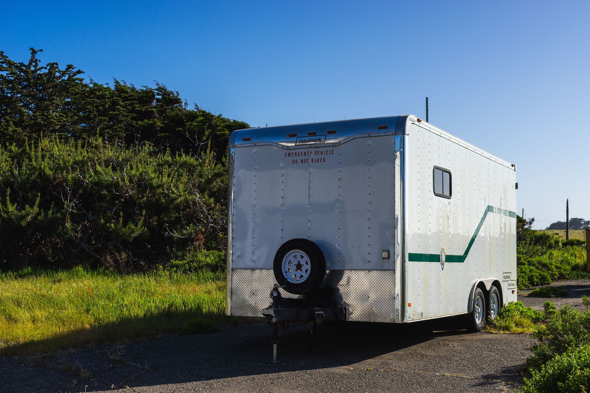 White utility trailer on a roadside, with a spare tire on the back and green trim, beside grass and trees under blue sky