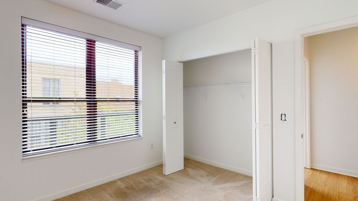 Here at Vim + Vigor Lofts in Wisconsin. A bright bedroom features soft carpeting, a large window, open closet doors, and a clean, spacious layout.