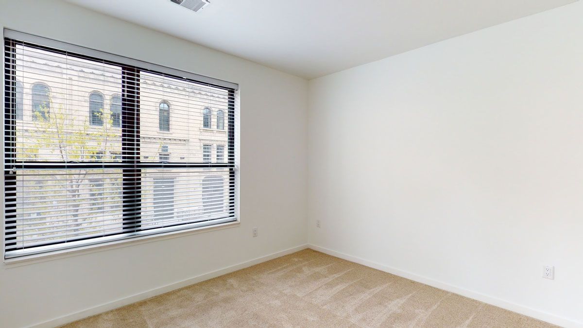Here at Vim + Vigor Lofts in Wisconsin. A bright empty bedroom features soft carpeting, large windows, clean white walls, and a spacious, welcoming layout.