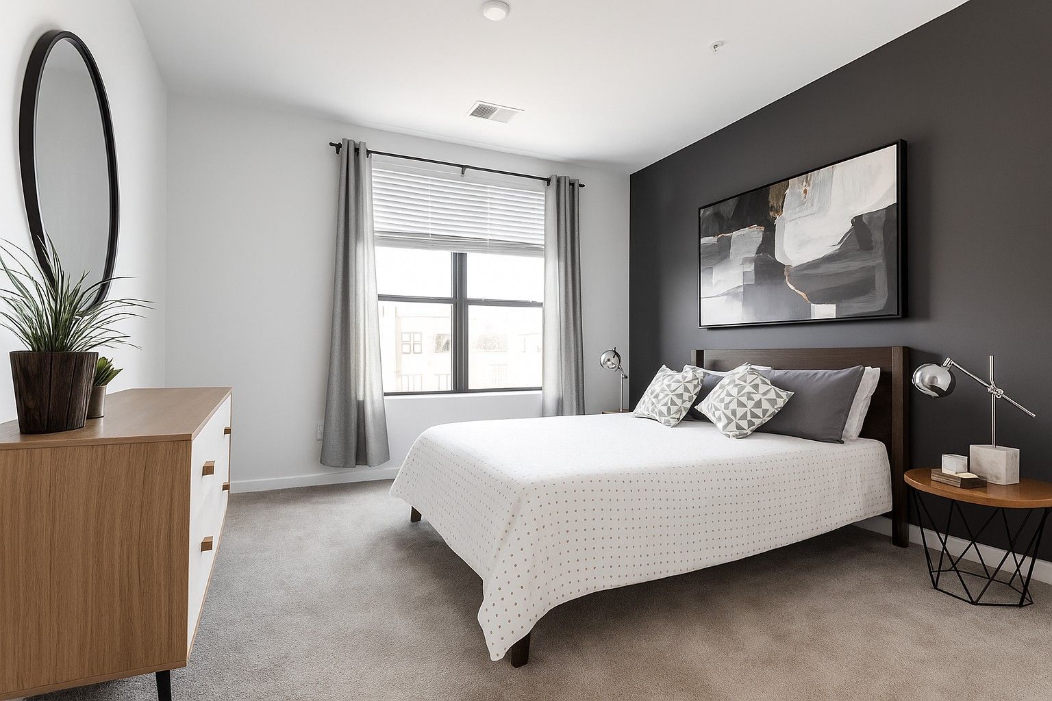 Here at Vim + Vigor Lofts in Wisconsin. A cozy bedroom features a dark accent wall, neutral bedding, soft carpeting, modern artwork, gray curtains, and warm wood furniture.