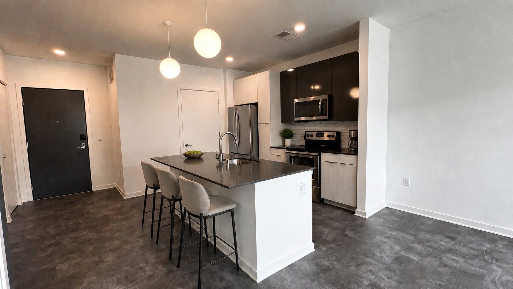 Here at Vim + Vigor Lofts in Wisconsin. A modern kitchen features a spacious island with bar seating, dark counters, stainless steel appliances, pendant lighting, and sleek two-tone cabinetry.