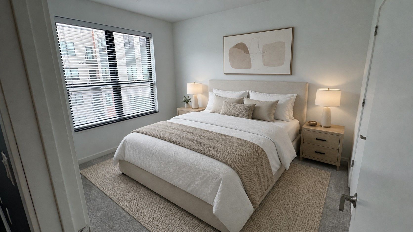 Here at Vim + Vigor Lofts in Wisconsin. A bright bedroom features neutral bedding, warm lamps, large windows, soft textures, and a clean, modern layout.