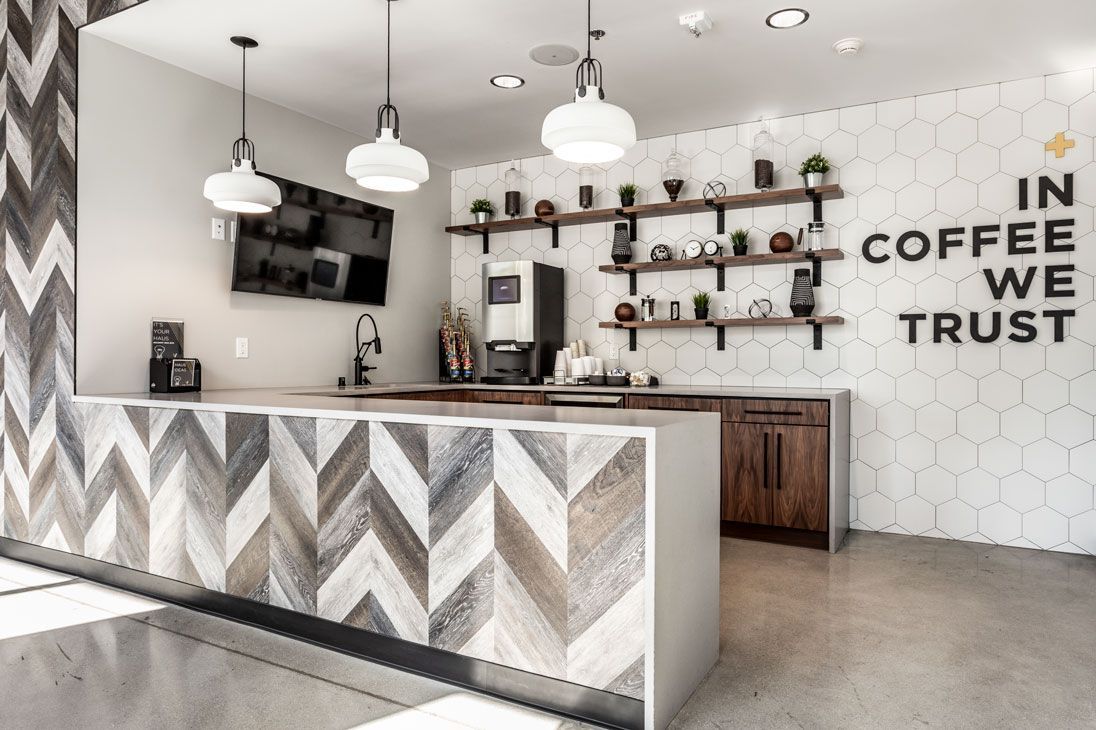 Here at Vim + Vigor Lofts in Wisconsin. A stylish coffee bar features patterned tile, warm wood cabinetry, open shelving, pendant lighting, and modern community seating nearby.