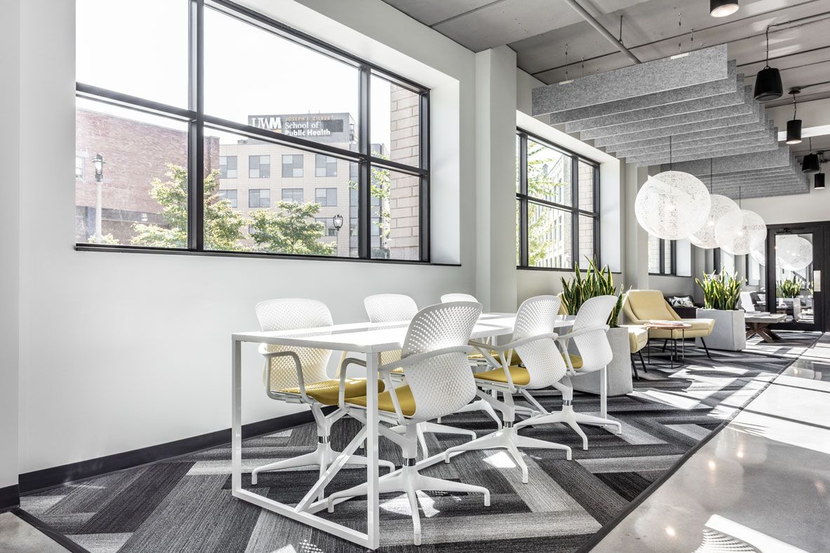 Here at Vim + Vigor Lofts in Wisconsin. A bright community workspace features white chairs, yellow seat accents, large windows, modern lighting, and a spacious shared table.
