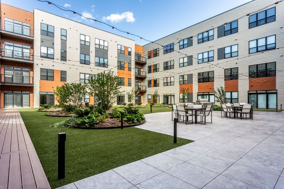 Here at Vim + Vigor Lofts in Wisconsin. A spacious courtyard features outdoor seating, string lights, landscaped greenery, modern apartment balconies, and an inviting patio area.