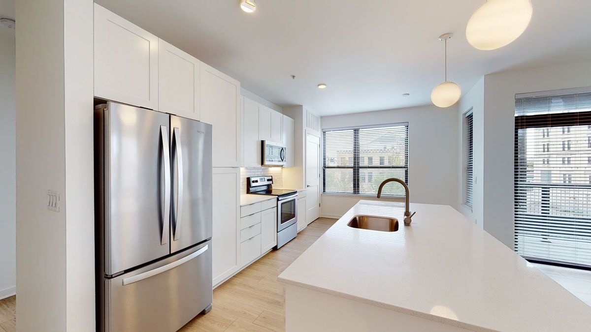 Here at Vim + Vigor Lofts in Wisconsin. A modern kitchen features stainless steel appliances, white cabinetry, a spacious island, warm flooring, and bright natural light.