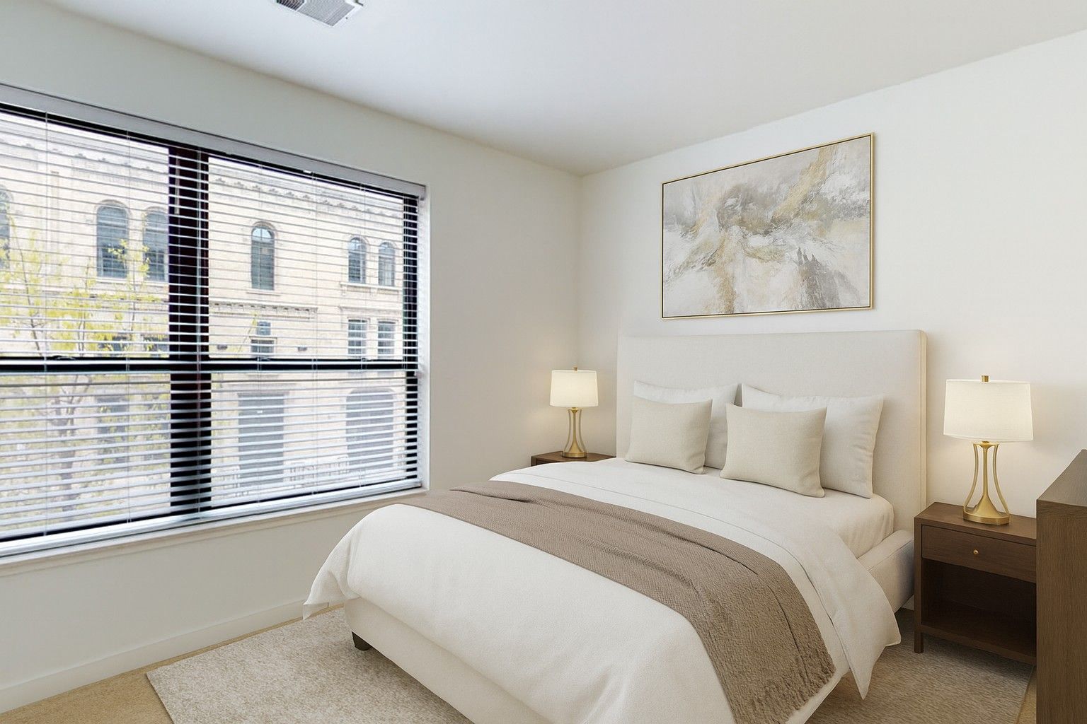 Here at Vim + Vigor Lofts in Wisconsin. A cozy bedroom features neutral bedding, warm lamps, soft carpeting, large windows, and modern wall art.