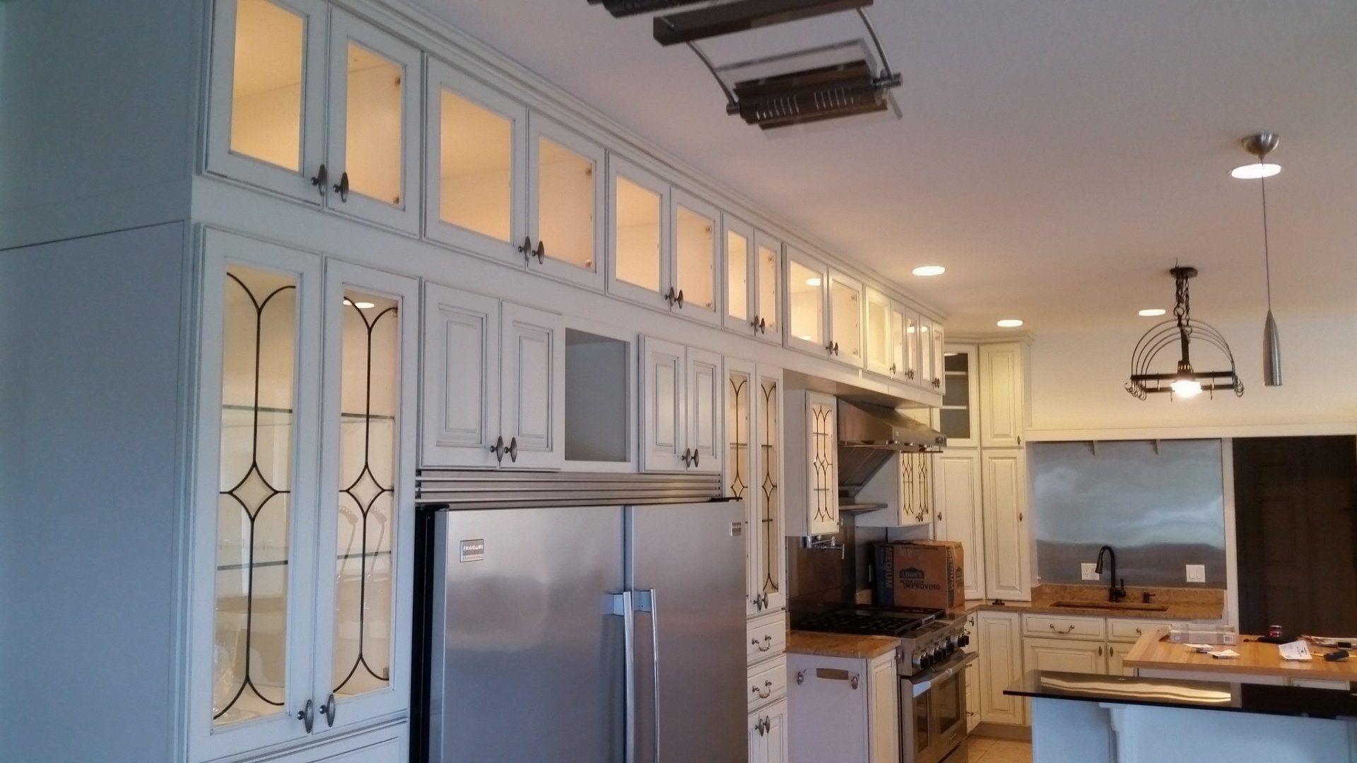 Cream-colored kitchen cabinets, including glass-fronted ones, surround a stainless steel refrigerator. Overhead lights illuminate the cabinets.