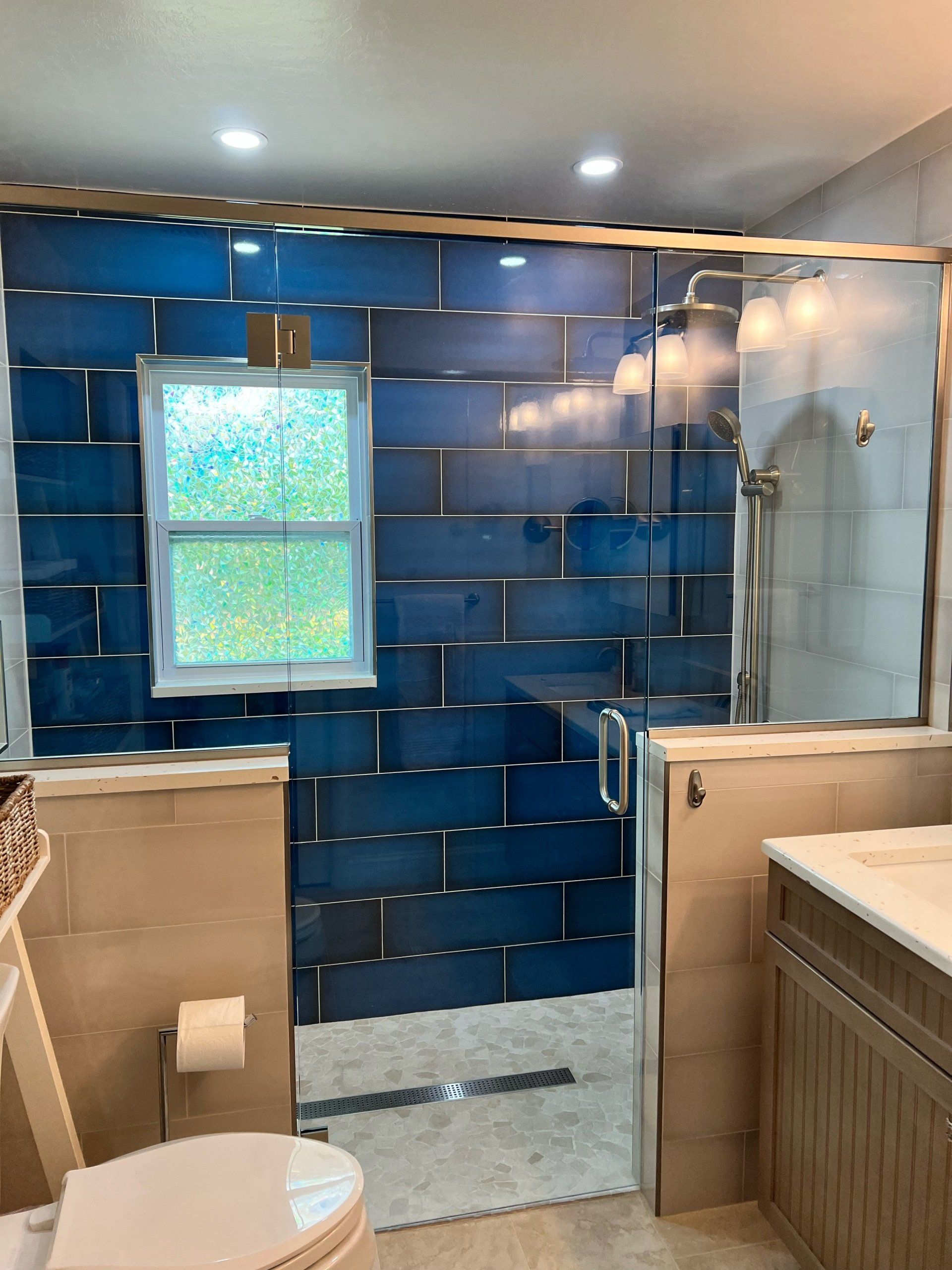 Blue tile shower with glass door, window, and vanity in a renovated bathroom.