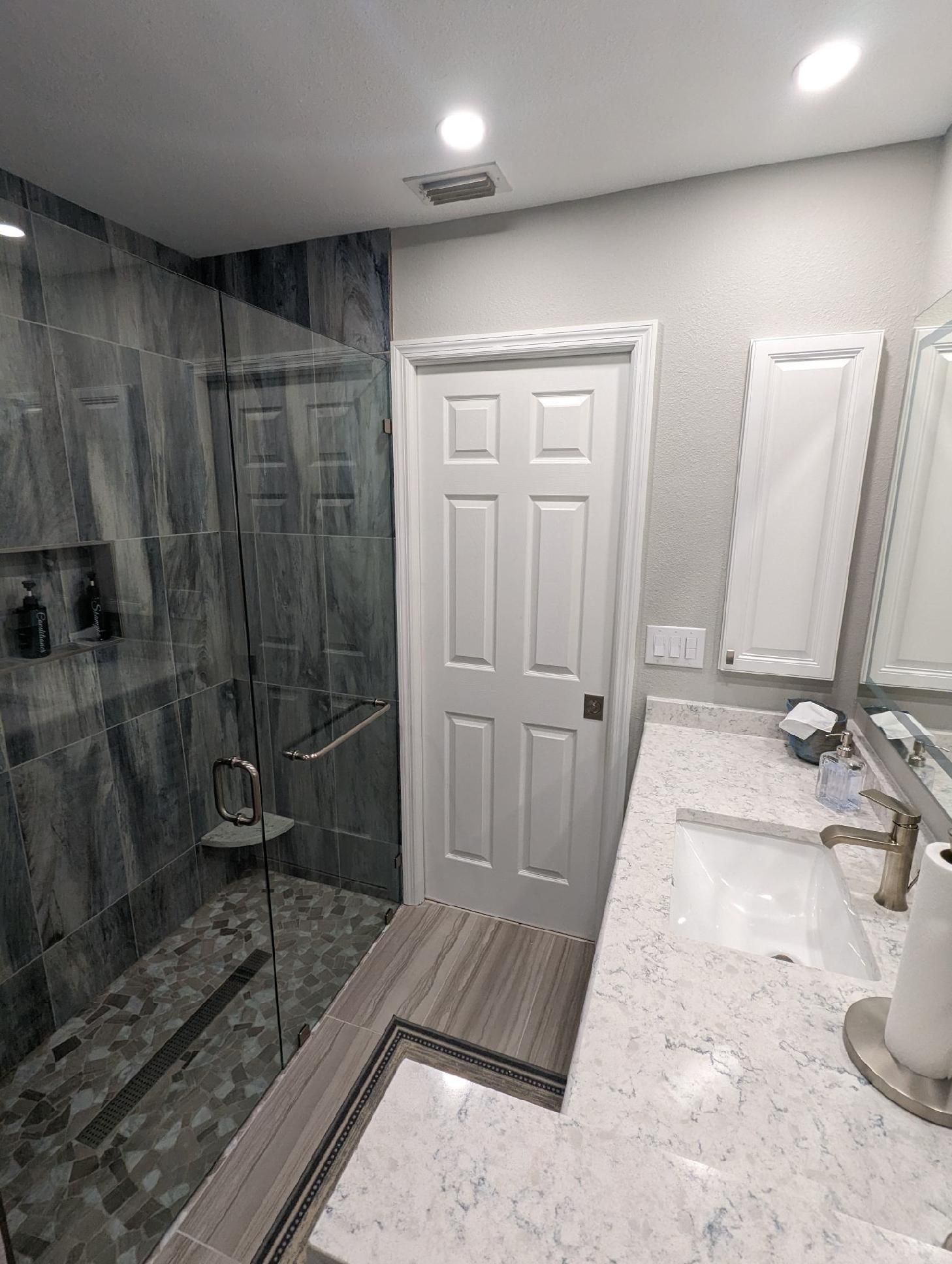 Bathroom with glass shower, white door, gray and white countertop, and large mirror.