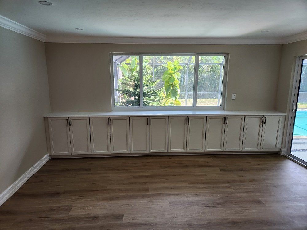 A room with a long white cabinet under a window, light brown floor, and a view of trees and a pool.