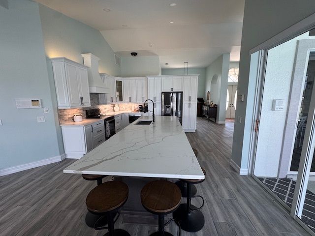 Spacious kitchen with white cabinets, marble island, and dark bar stools. Gray wood-like floors, high ceilings, and light blue walls.