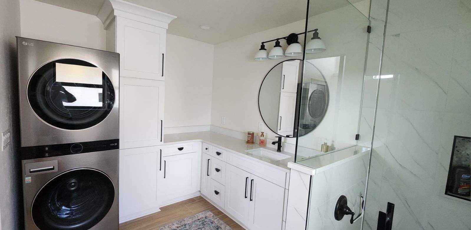 A modern bathroom with a washer/dryer, white cabinets, a circular mirror, and a glass shower.