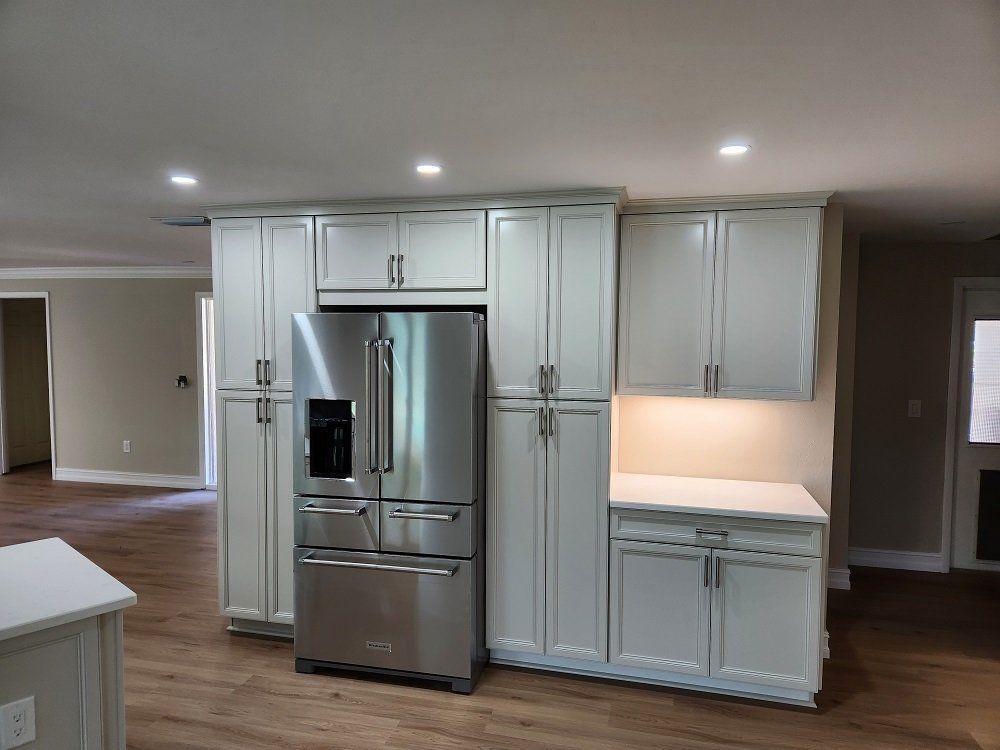 White kitchen cabinets surround a stainless steel refrigerator. A countertop with under cabinet lighting is visible.