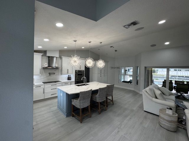 Spacious kitchen with white cabinets, marble island, and dark bar stools. Gray wood-like floors, high ceilings, and light blue walls.