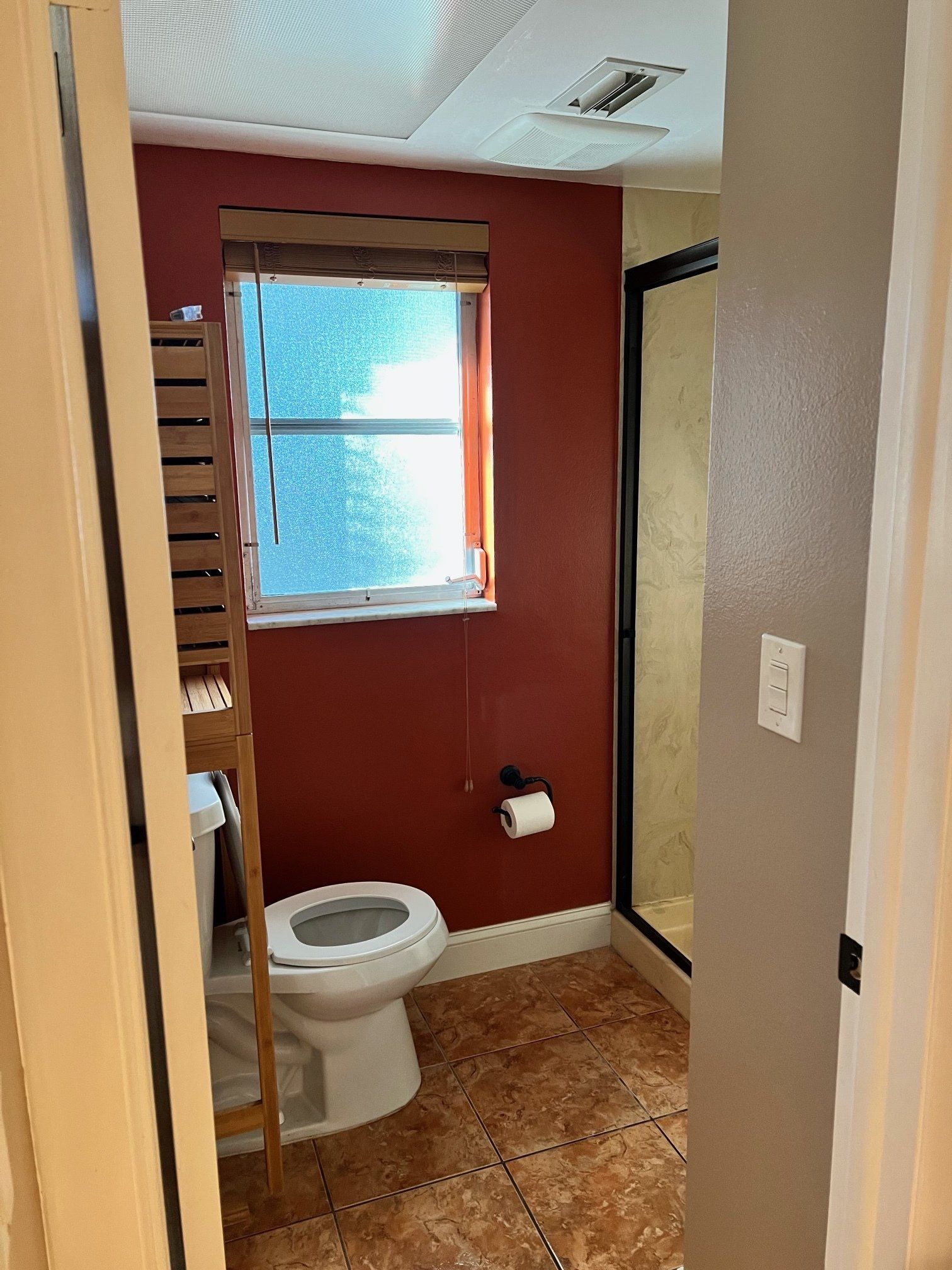 Bathroom with red wall, window, toilet, shower, and shelving unit.