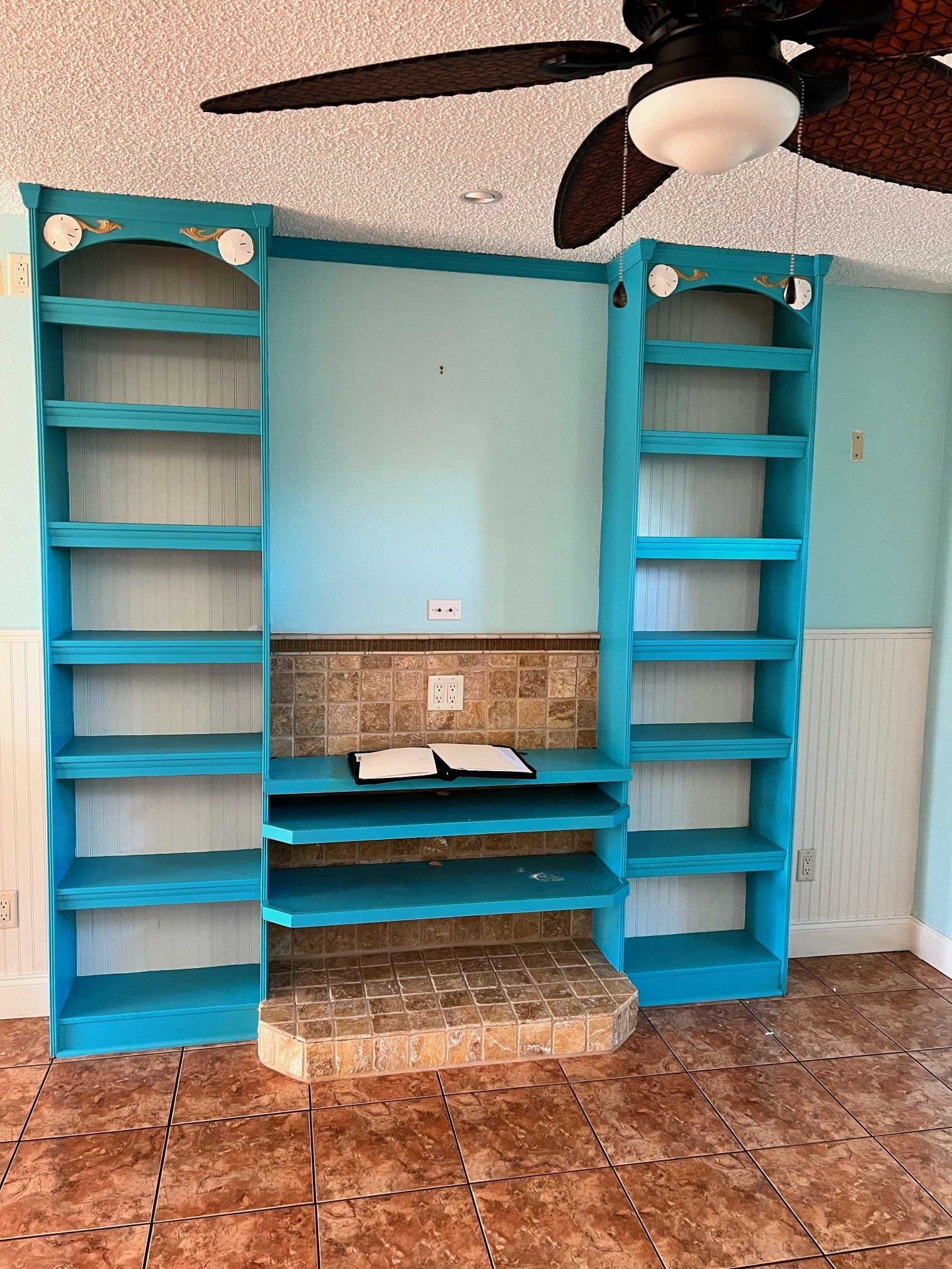 Built-in turquoise bookshelf unit with multiple shelves, center desk area, on tile floor. Ceiling fan overhead.