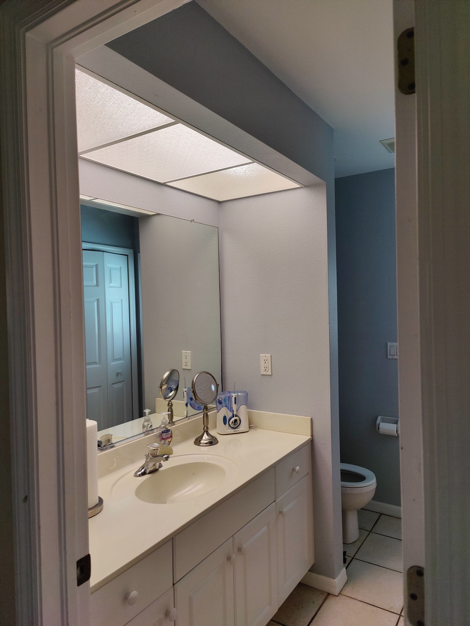 Bathroom interior with white vanity, mirror, and blue walls. Doorway on right, toilet on right.