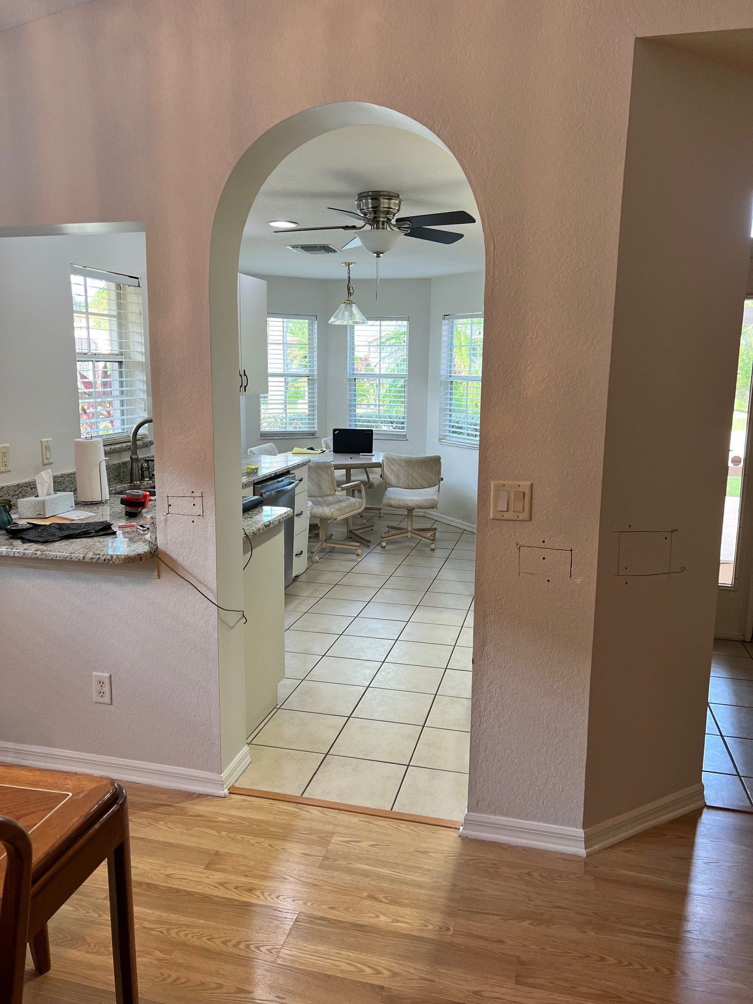 An arched doorway leads to a brightly lit room with windows, tile flooring, and a ceiling fan.
