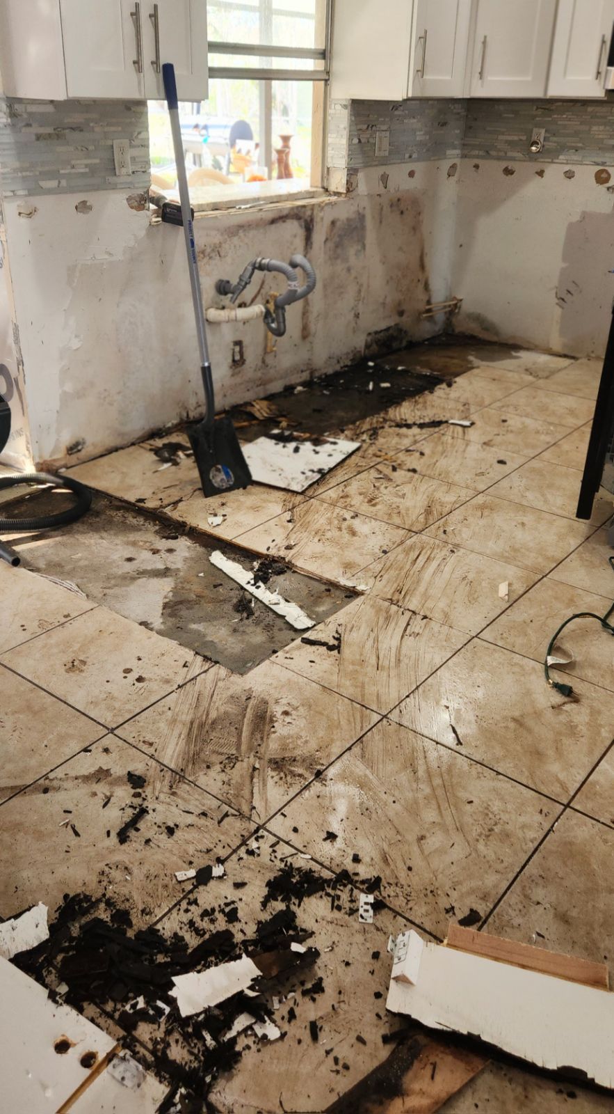 Kitchen demolition: tiles removed, debris scattered, walls exposed, cabinets remain.