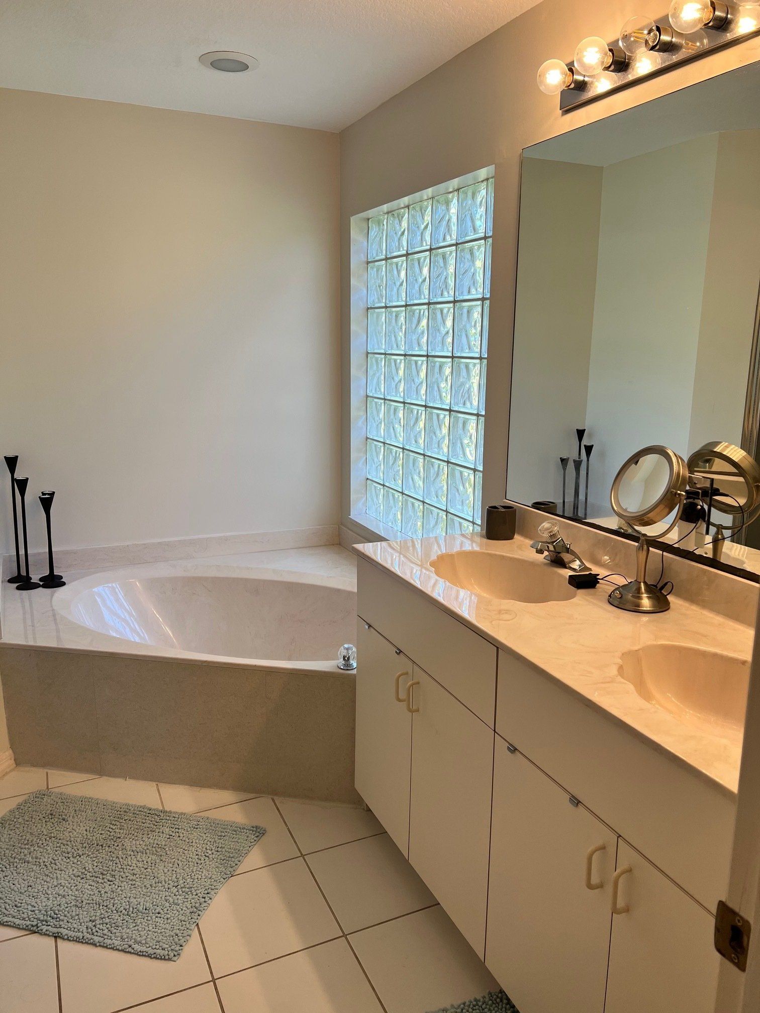 Bathroom with a built-in tub, double sink vanity, and glass block window. Light blue rug on tile floor.