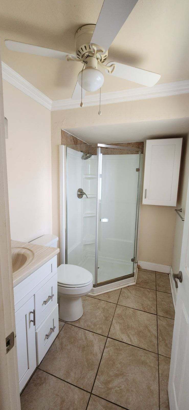 Bathroom with toilet, shower, vanity, and ceiling fan. Beige walls and tile.
