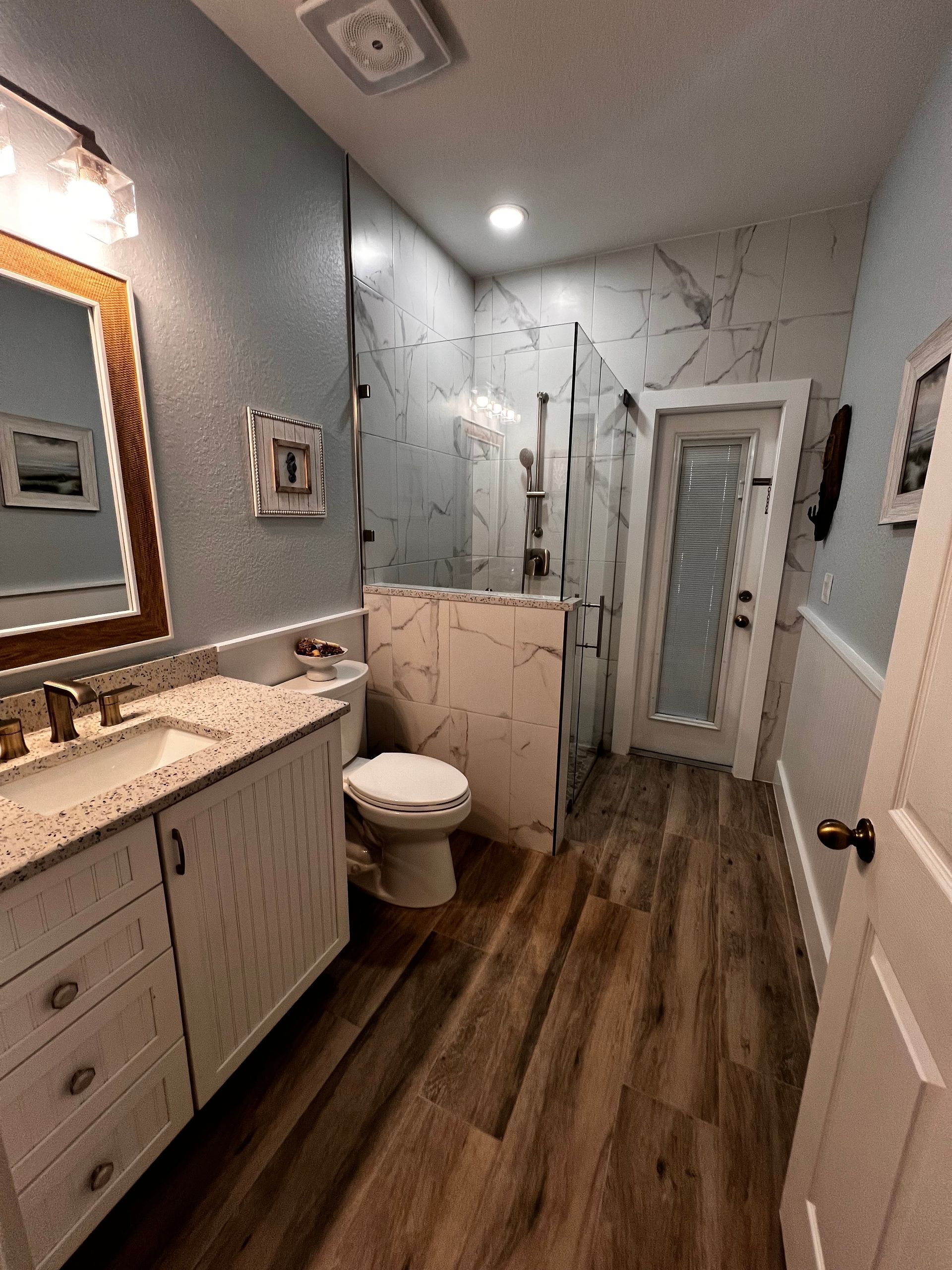 Bathroom with white vanity, toilet, and shower, featuring wood-look flooring and marble-patterned wall tiles.