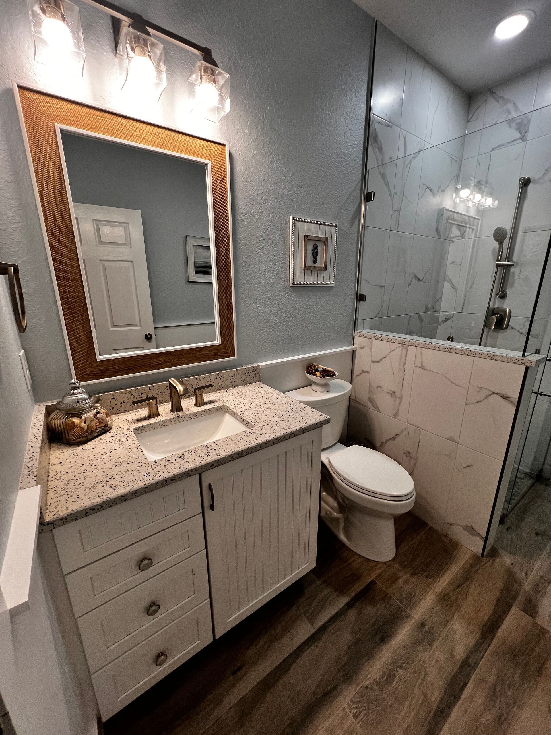 Bathroom with white vanity, toilet, marble-tiled shower, and framed mirror. Wooden floor and blue-gray wall.