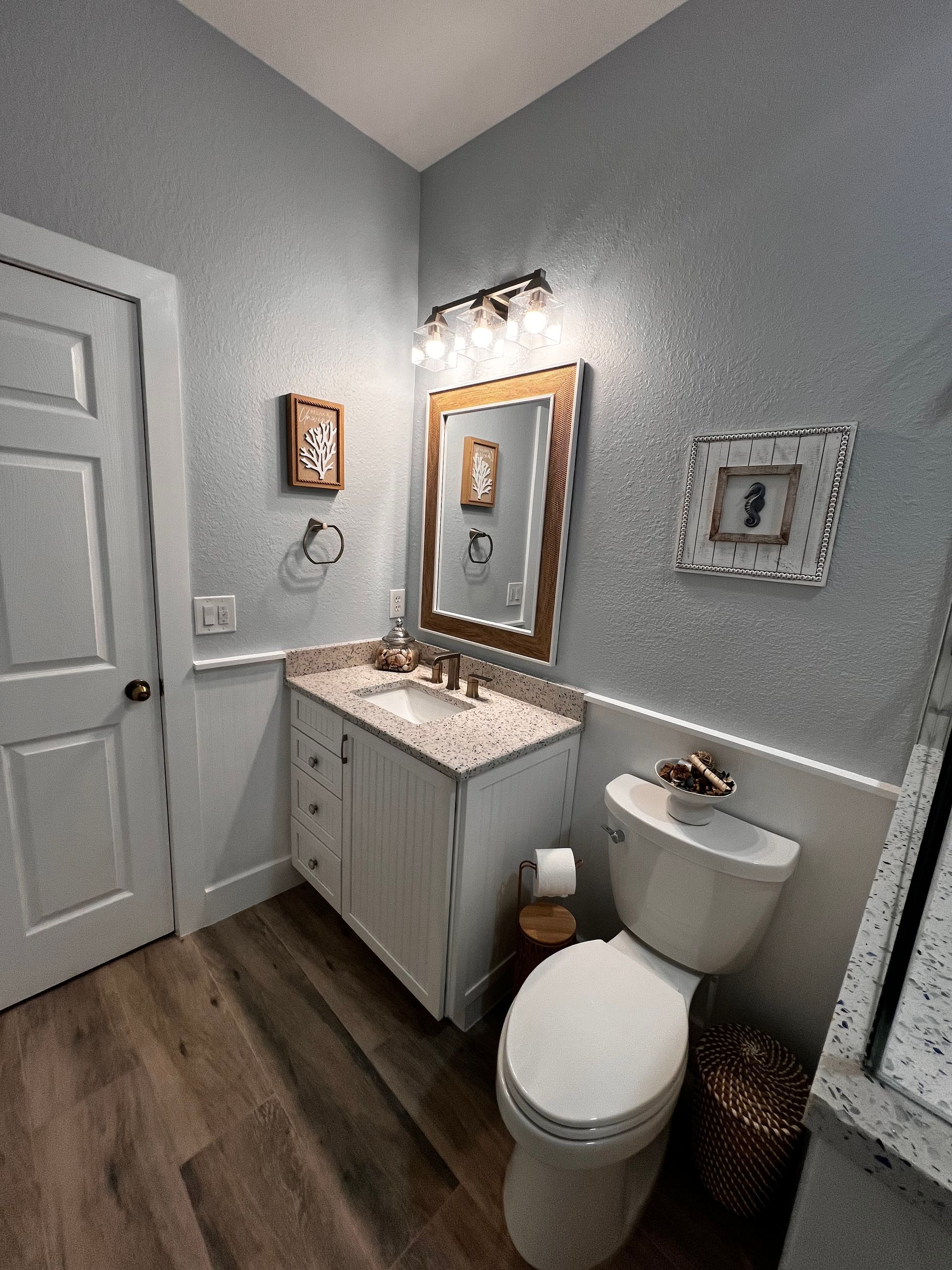 Small bathroom with a toilet, vanity, and wooden floor. Light blue walls, white trim, and a decorative mirror.