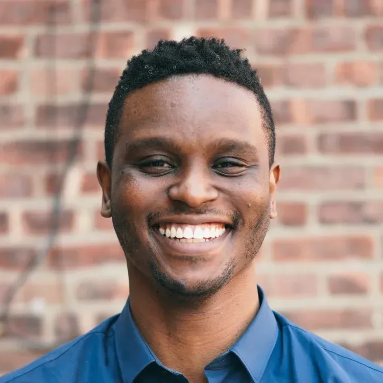 Man with dark skin smiling in front of a brick wall, wearing a blue shirt.