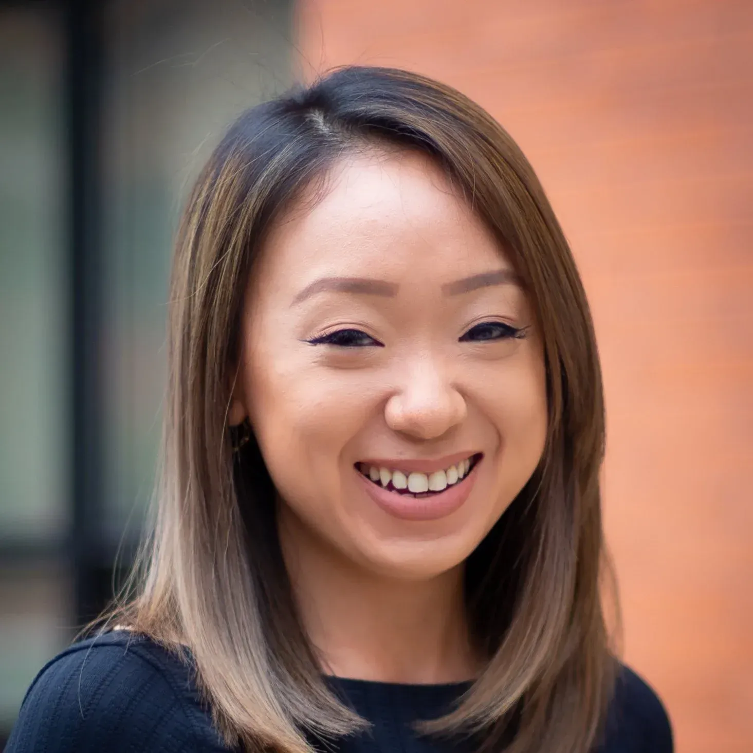 Woman with a broad smile, light brown hair, black top, against a brick wall background.