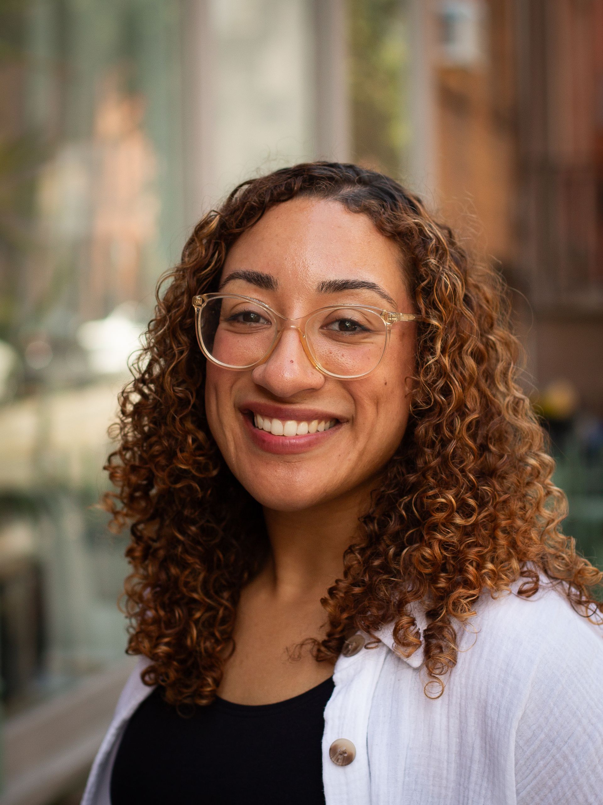 Smiling person with curly hair and glasses in a light top, standing indoors against a blurred background
