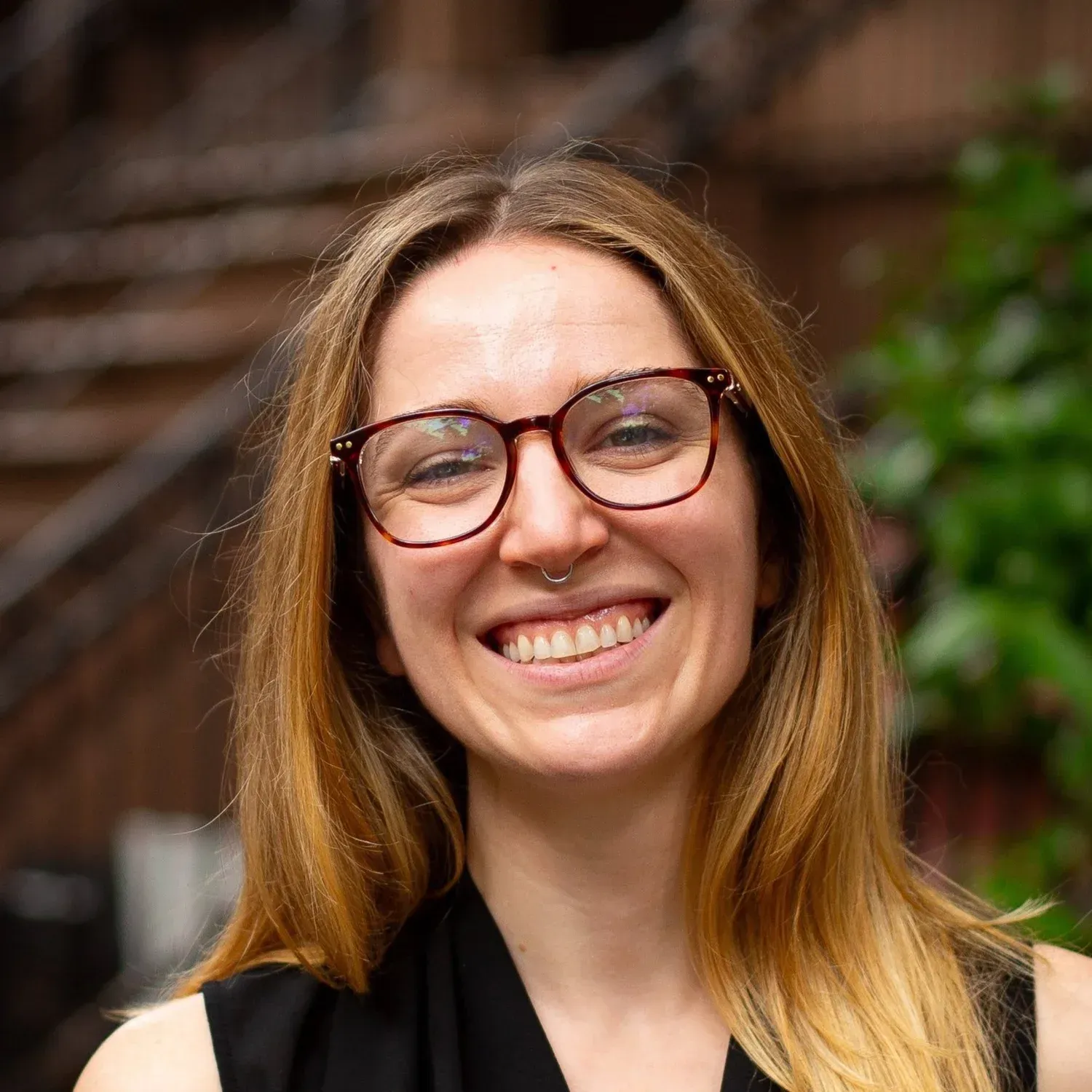 Woman with glasses and nose ring smiles broadly outdoors.