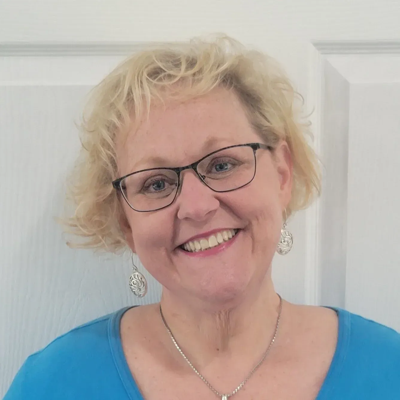 Woman with glasses smiles; blonde hair, blue shirt, silver earrings, and necklace.