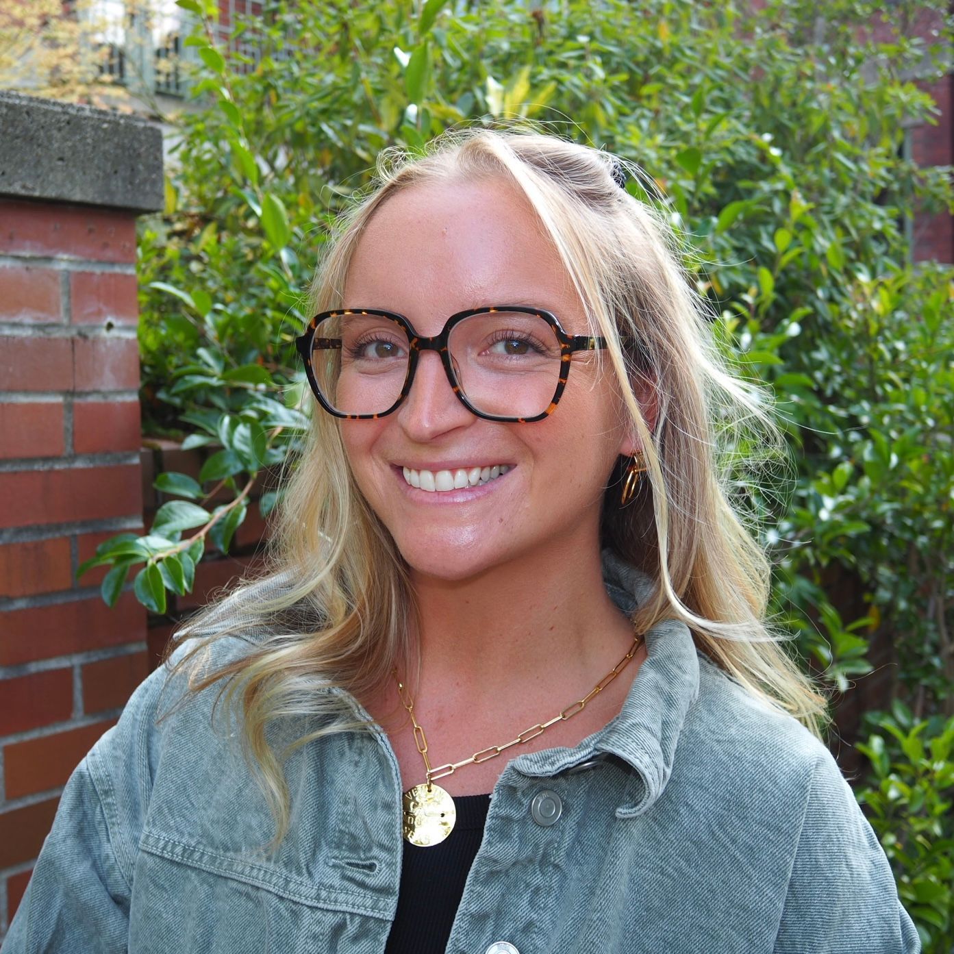 Smiling person with glasses and blonde hair in a denim jacket standing outdoors by greenery.