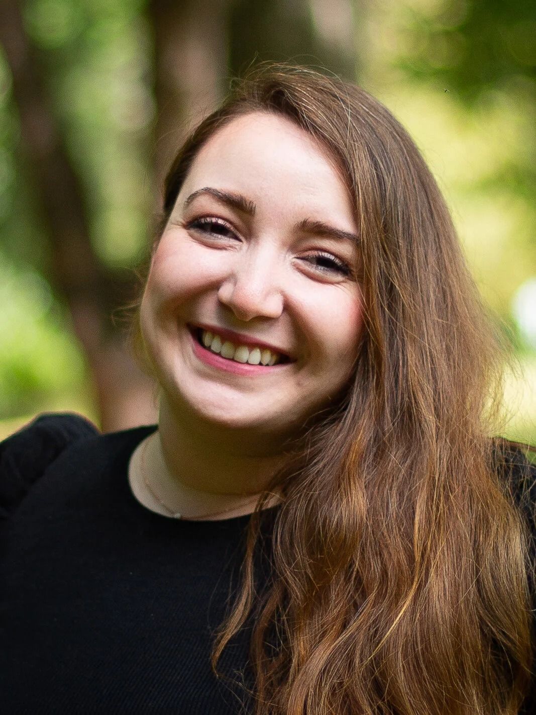 Woman with long brown hair, smiling, outdoors. Wearing a black top, blurred green background.