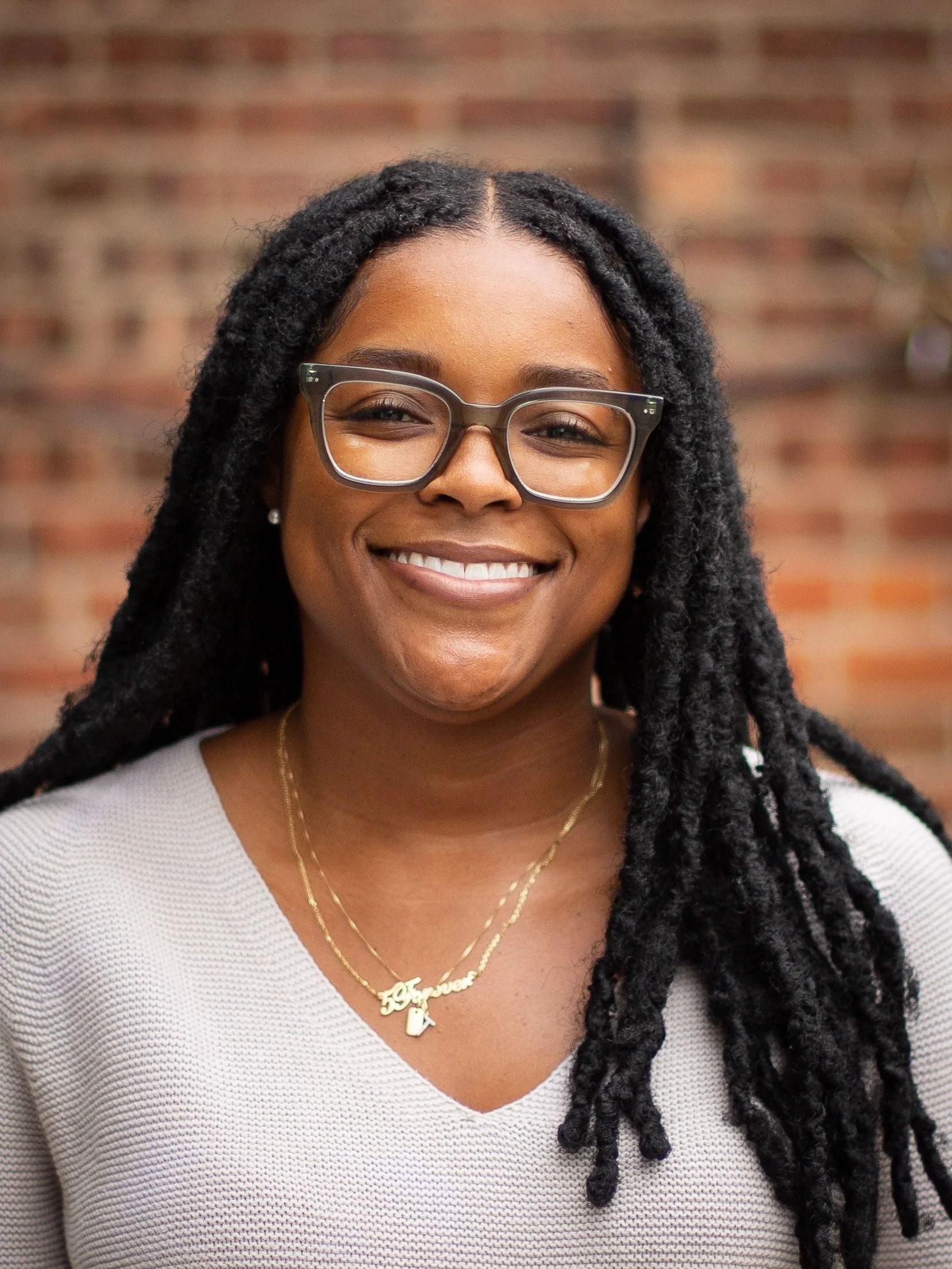 Woman with locs and glasses smiles in front of a brick wall. She wears a cream sweater and a gold necklace.