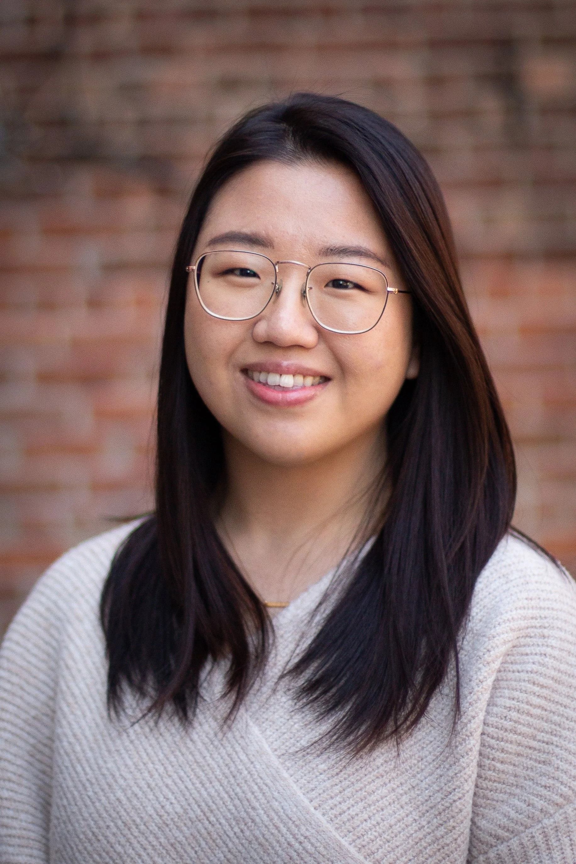 Woman with glasses smiles in front of a brick wall.