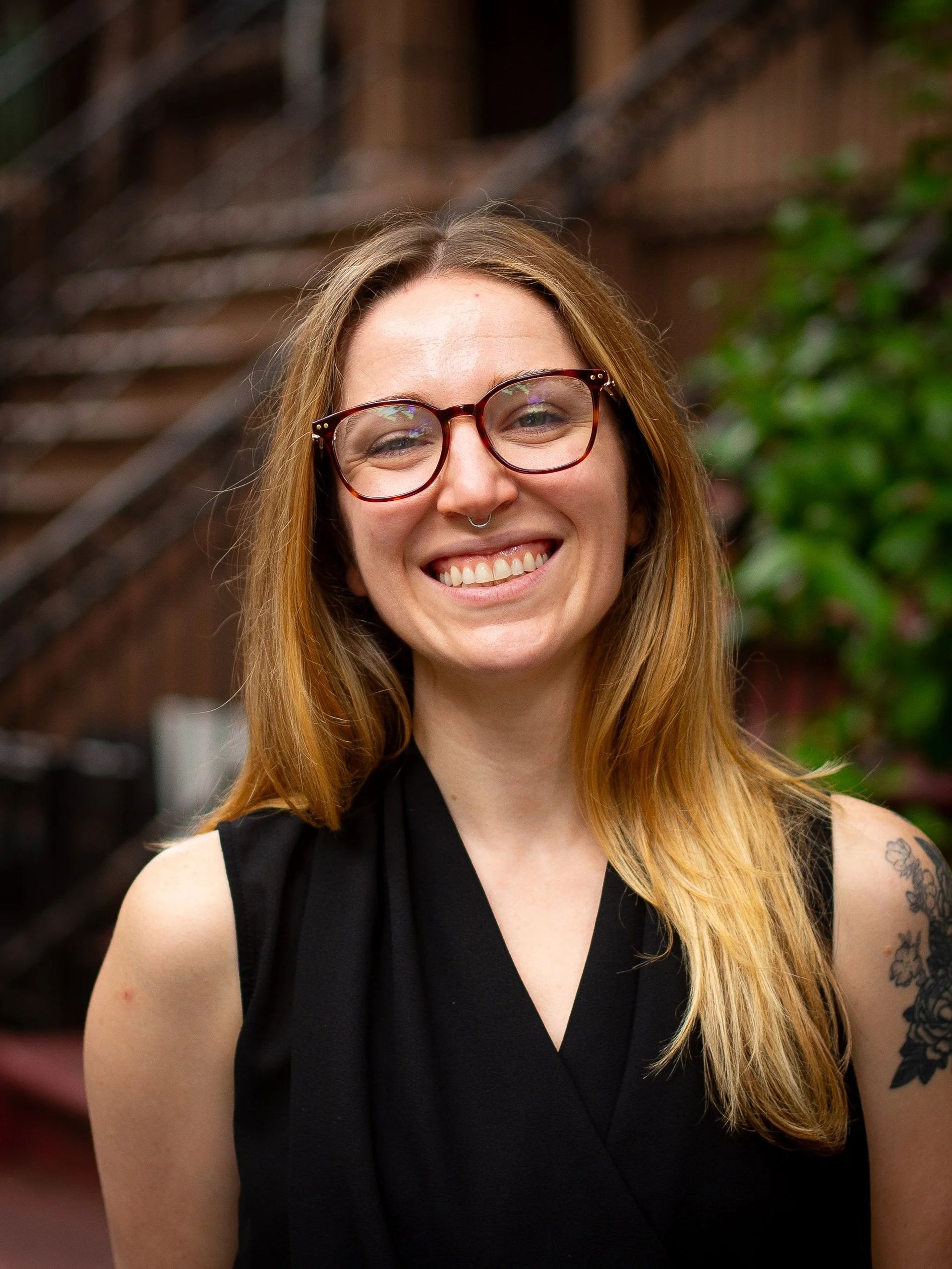 Woman with glasses smiling, wearing a black sleeveless top, outdoors in front of a brick building.