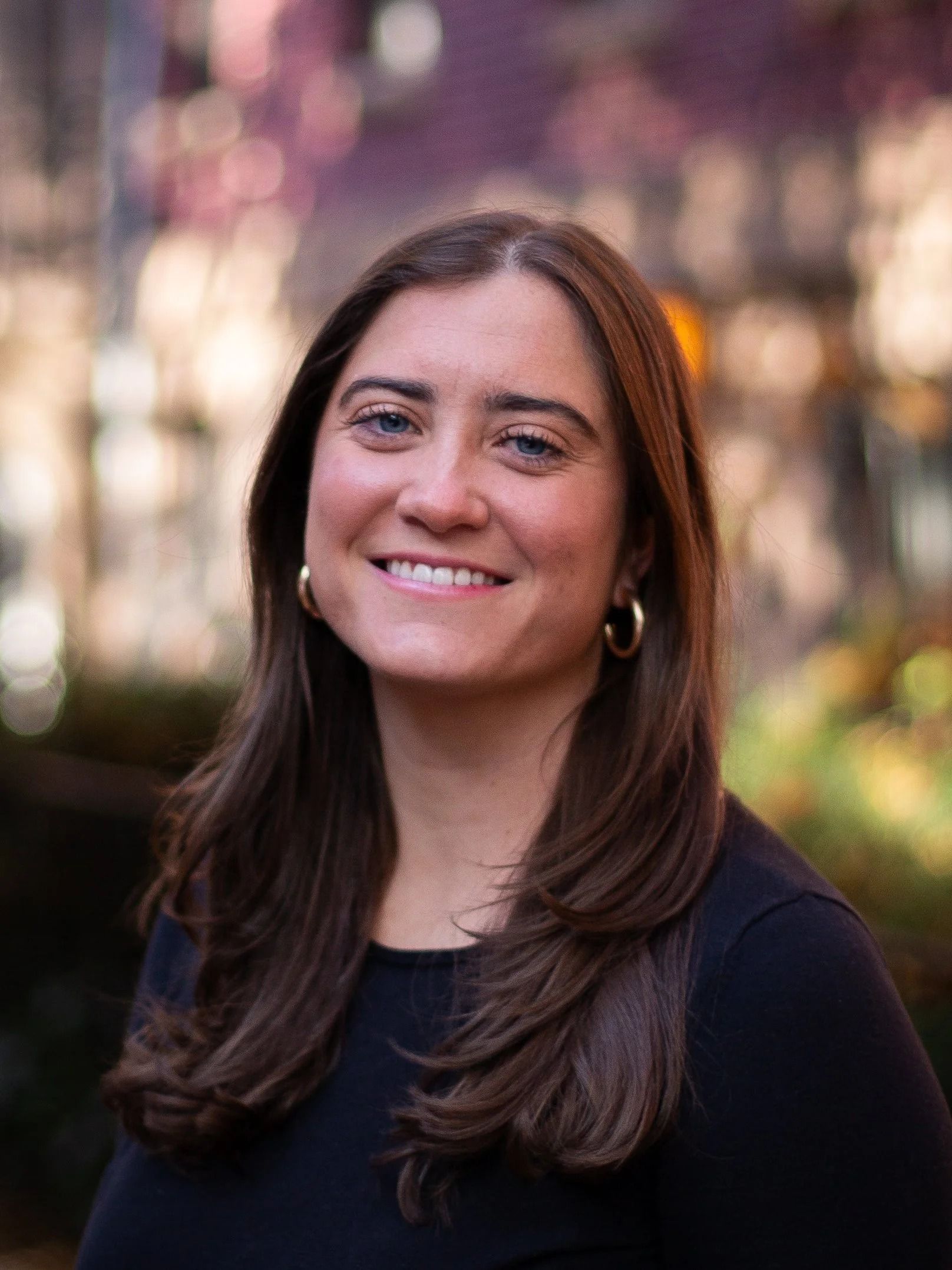 Woman with long brown hair smiles, wearing gold hoop earrings, a black shirt, outdoors with blurred background.