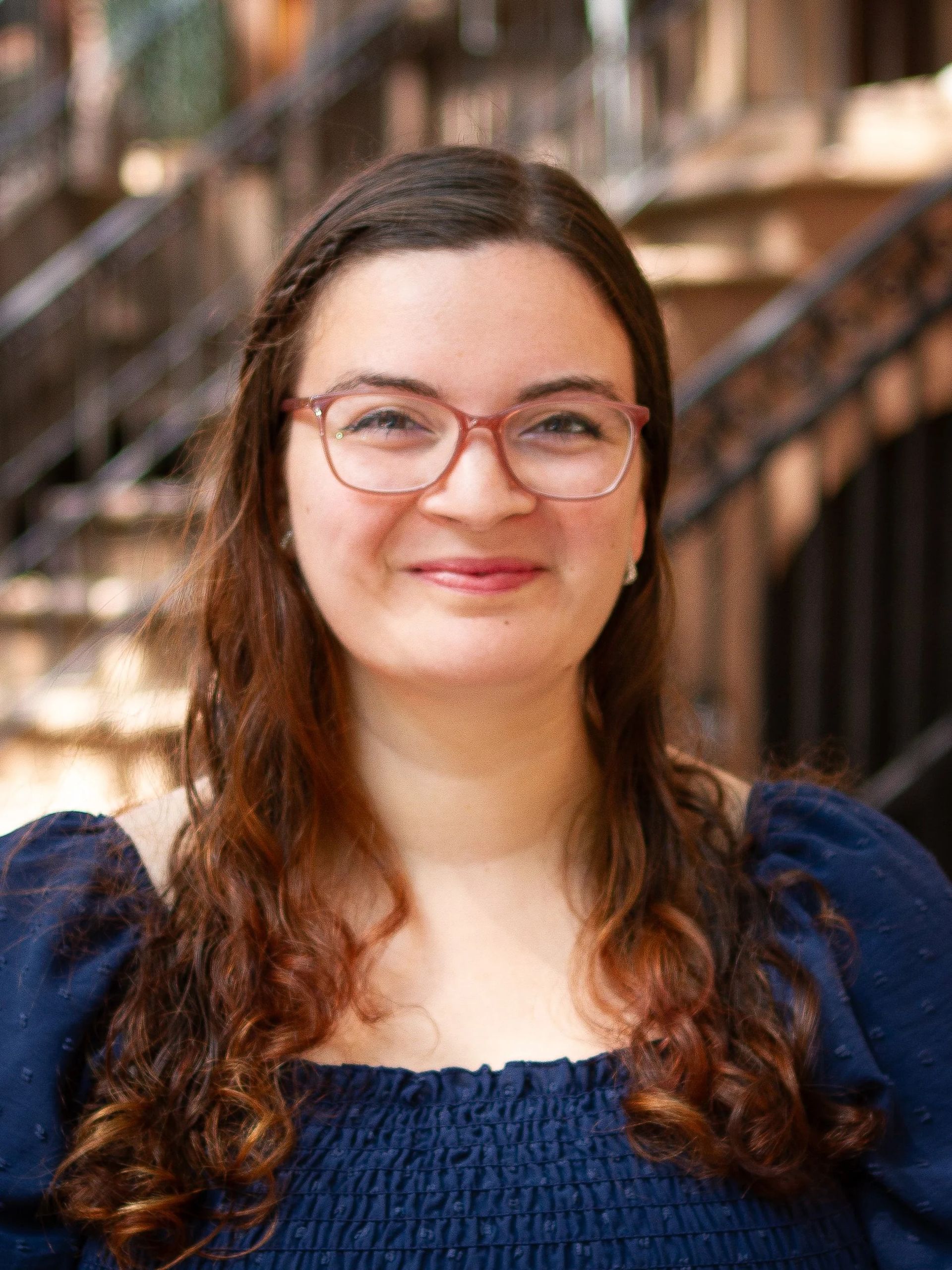 Woman with glasses smiles, looking forward. Wears a dark blue top and stands near a staircase in a building.